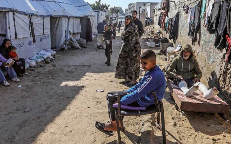 Geflüchtete Palästinenser aus dem Gazastreifen sitzen und stehen auf einer Lehmstraße. Links sind Zelte zu sehen, rechts aufgehängte Wäsche an einer Mauer.