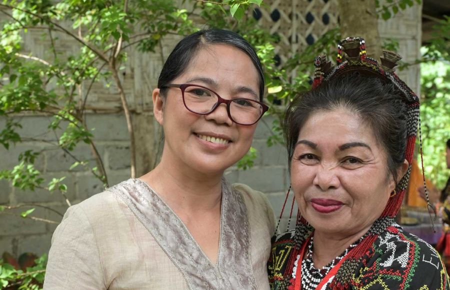 Zwei Frauen aus Mindanao/Philippinen lächeln vor einer Hauswand mit Bäumen in die Kamera: links Projektleiterin Jocelyn Aquiatan und rechts eine Begünstigte.