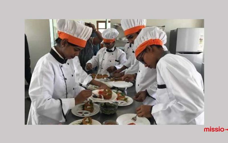 Vier ehemalige Straßenkinder stehen in weißer Kochuniform und Kochmützen in einer Küche und dekorieren Teller mit Essen.